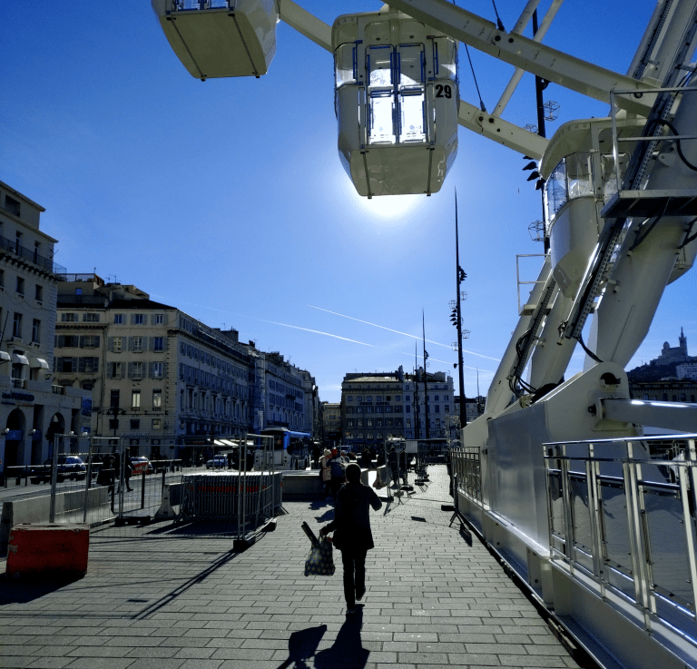 Vue sur le Vieux-Port de Marseille, protégé par l'ombre de la grande roue. Au loin, en haut sur la droite, la Bonne Mère qui veille sur le ciel bleu.